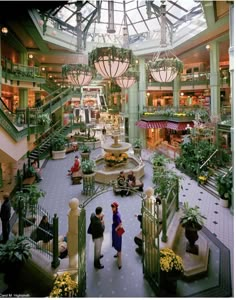 two people are walking through the atrium of a shopping mall with many plants and flowers