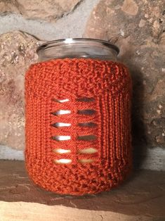 an orange crocheted mason jar cover sitting on top of a wooden table next to a stone wall