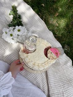 a person is holding a cake on a blanket with flowers in the grass behind it