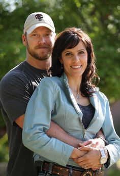 a man and woman standing next to each other in front of trees with their arms around each other