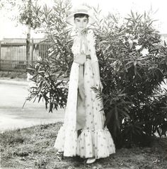 an old black and white photo of a woman standing in front of a bush wearing a hat