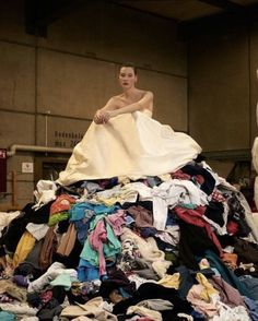 a woman sitting on top of a pile of clothes in an industrial building, surrounded by piles of shirts