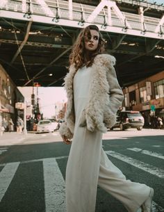 a woman is walking down the street wearing white pants and a fur coat with her hands in her pockets