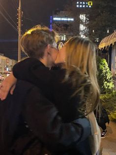 a man and woman kissing on the street at night with city lights in the background