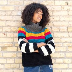 a woman standing in front of a brick wall wearing a multicolored striped sweater