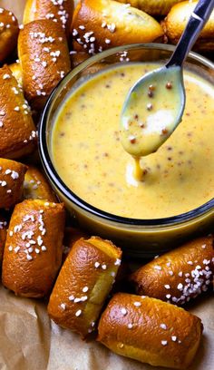 a spoon full of mustard sauce being held over some bread rolls with sesame seed sprinkles