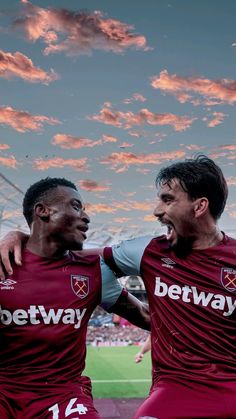 two men in maroon uniforms standing next to each other on a soccer field with the sky and clouds behind them