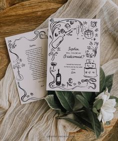 an open book on top of a wooden table next to a white flower and green leafy plant