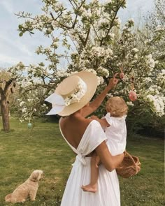 a woman in a white dress and straw hat holding a baby while standing next to an apple tree