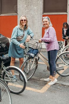 two women standing next to each other with bicycles in front of them on the street