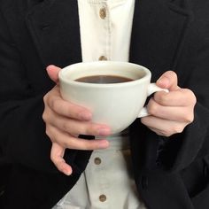 a woman holding a cup of coffee in her hands while wearing a black jacket and white shirt