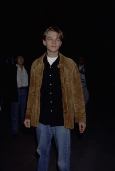 a young man standing in the dark with his hands in his pockets and looking at the camera