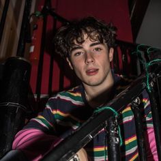 a young man with curly hair and striped shirt sitting in front of a stair railing