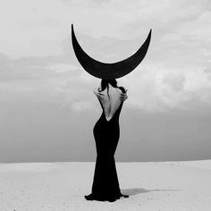 a woman in a long black dress with a crescent moon above her head on the beach