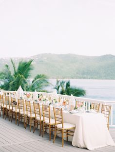 a long table set up with white linens and gold chairs on a deck overlooking the ocean