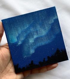 a hand holding up a small painting with an aurora bore in the sky behind it