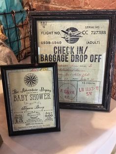 two framed baby shower signs sitting on top of a table next to a brick wall