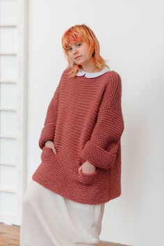 a woman standing in front of a white wall wearing a red sweater and white pants