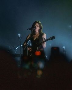 a woman holding a guitar while standing in front of microphones and singing on stage