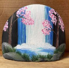 a painted rock with pink flowers on it and waterfall in the background, sitting on a wooden surface