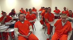 a group of men in orange prison uniforms sitting at desks