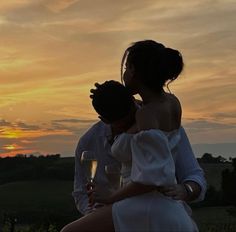 a man and woman sitting on top of each other holding glasses of wine at sunset