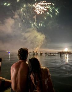 two people sitting on a dock watching fireworks go off in the sky