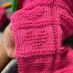 a person holding up a pink knitted sweater in their left hand and knitting needles in the background