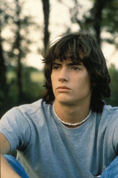 a young man with long hair wearing a gray shirt sitting in front of some trees