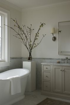 a white bath tub sitting under a window next to a sink and a vase with flowers