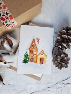 a card with a watercolor painting of a house on it next to pine cones