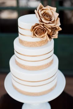 a white and gold wedding cake with three roses on the top, sitting on a table