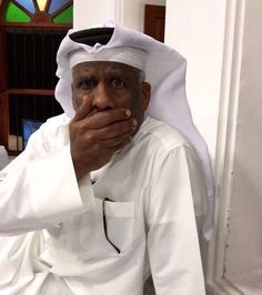 a man sitting down with his hand on his face and looking at the camera while wearing a white outfit