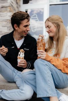 a young man and woman sitting on the ground eating ice cream cones while looking at each other