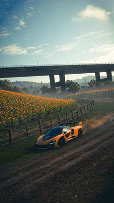 an orange sports car driving down a dirt road next to a field of yellow flowers