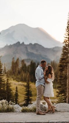 a man and woman standing next to each other in front of mountains with pine trees