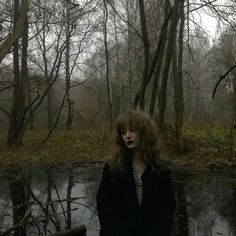 a woman with long hair standing in front of a body of water surrounded by trees