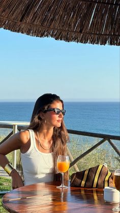 a woman sitting at a table with an orange juice in front of her and the ocean behind her