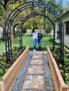 two women are standing under an arch in a garden with plants and stones on the ground