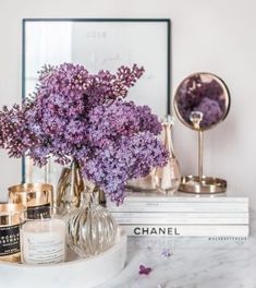 purple flowers in a glass vase on a marble counter top next to books and candles