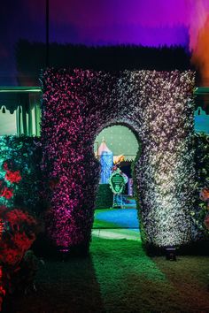 an archway in the middle of a garden with flowers growing around it at night time