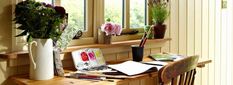 a wooden desk topped with lots of flowers and art supplies next to a large window