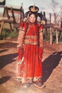 an old photo of a woman in red dress and headdress standing on dirt ground