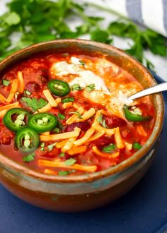 a close up of a bowl of chili soup with cheese and green peppers on the side