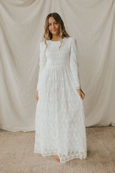 a woman standing in front of a white backdrop wearing a long sleeved wedding dress