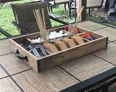 an outdoor picnic table with drinks, snacks and utensils in a wooden tray