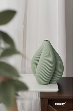 a green vase sitting on top of a wooden table next to a white window curtain