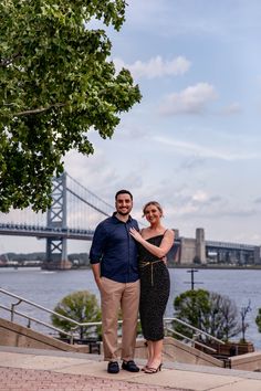 a man and woman standing next to each other in front of a river with a bridge in the background