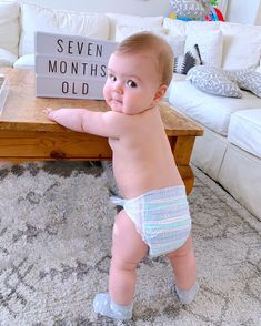 a baby standing in front of a coffee table with a sign on it that says seven months old