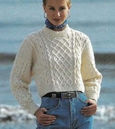 a woman standing next to the ocean wearing a white sweater and blue jeans with her hands on her hips